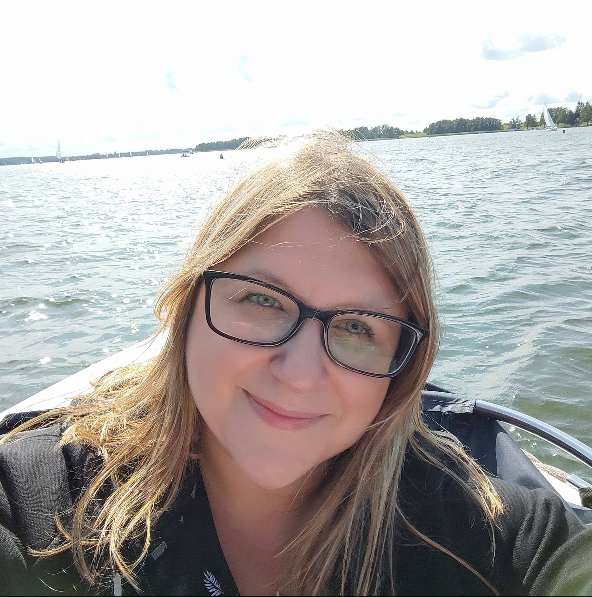 Uśmiechnięta kobieta w okularach na łodzi, w tle jezioro z żaglówkami i zielony brzeg.