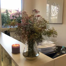 Recepcja z bukietem kolorowych gips&oacute;wek w szklanym wazonie ze złotą podstawą, zapaloną fioletową świecą i logo 'SATIS' na ścianie