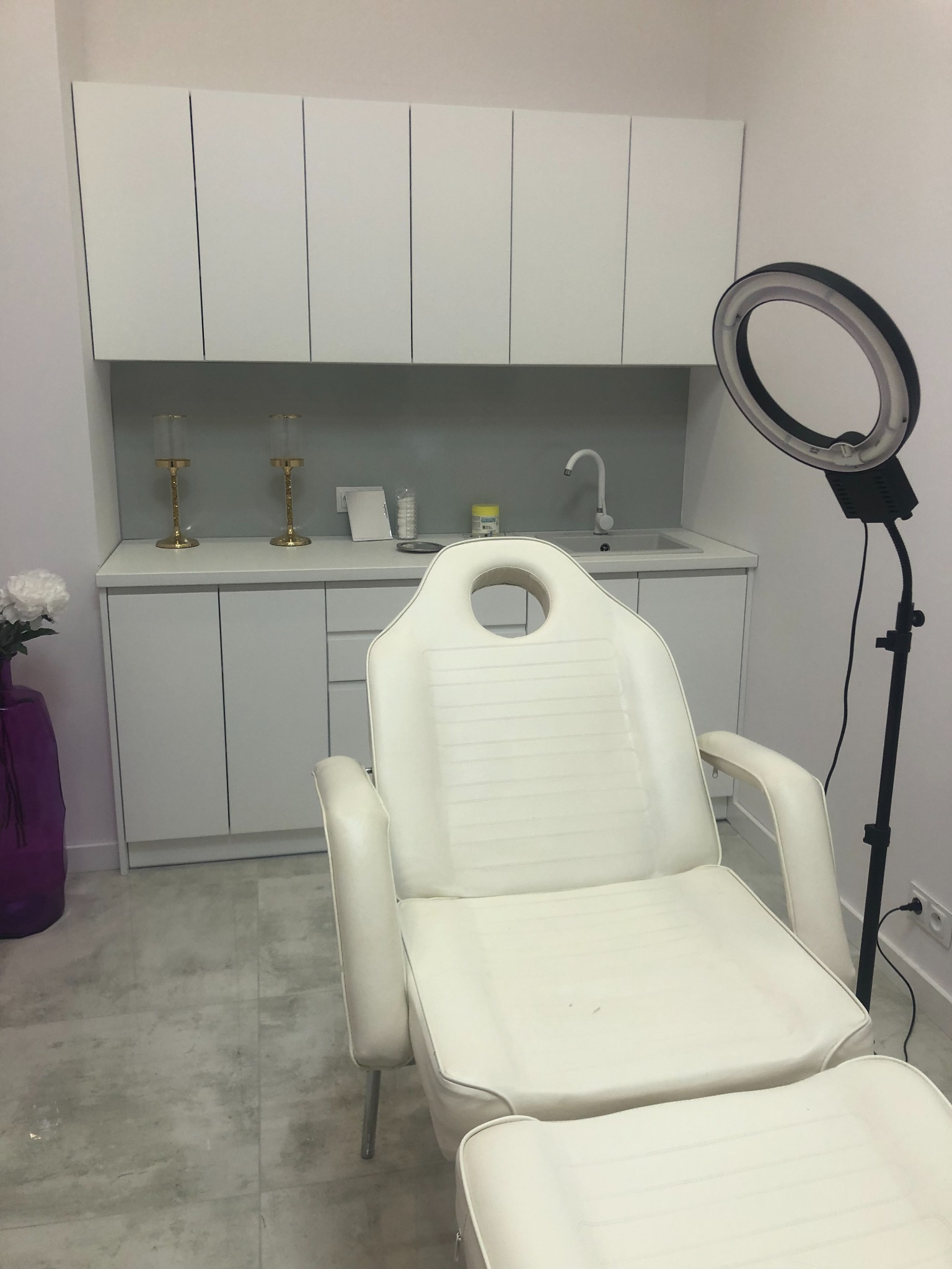 Jasne wnętrze gabinetu kosmetycznego z białym fotelem zabiegowym z podnóżkiem, lampą pierścieniową, szafkami i umywalką.
