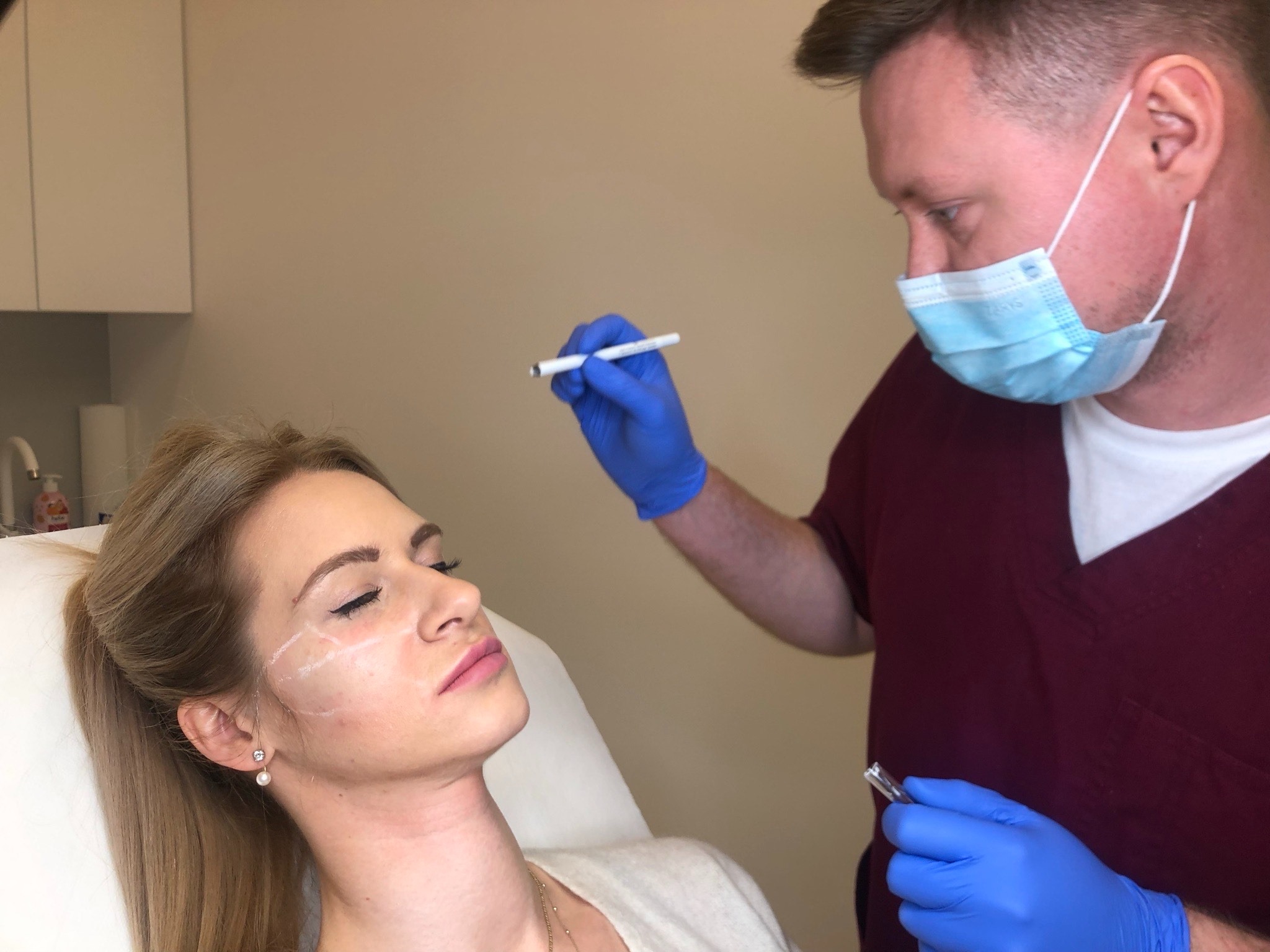 Lekarz w masce i rękawiczkach zaznacza markerem obszary na twarzy kobiety leżącej na leżance, przygotowując ją do zabiegu.