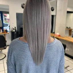 Tył głowy kobiety z prostymi, popielatymi włosami, ostrzyżonymi w szpic, widoczne w salonie z jasnymi ścianami i meblami.