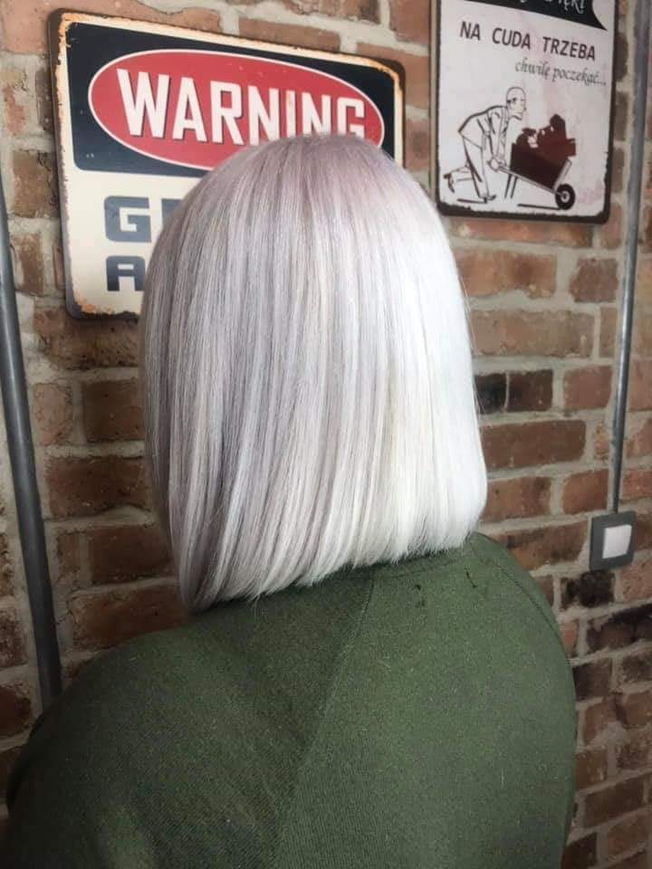 Tył głowy kobiety z krótką, platynową fryzurą typu bob, na tle ceglanej ściany z dekoracyjnymi tabliczkami.