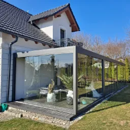 Szklana zabudowa tarasu z metalową ramą, przylegająca do białego domu z ciemnym dachem. Wewnątrz rośliny doniczkowe i meble ogrodowe. Słoneczny dzień.