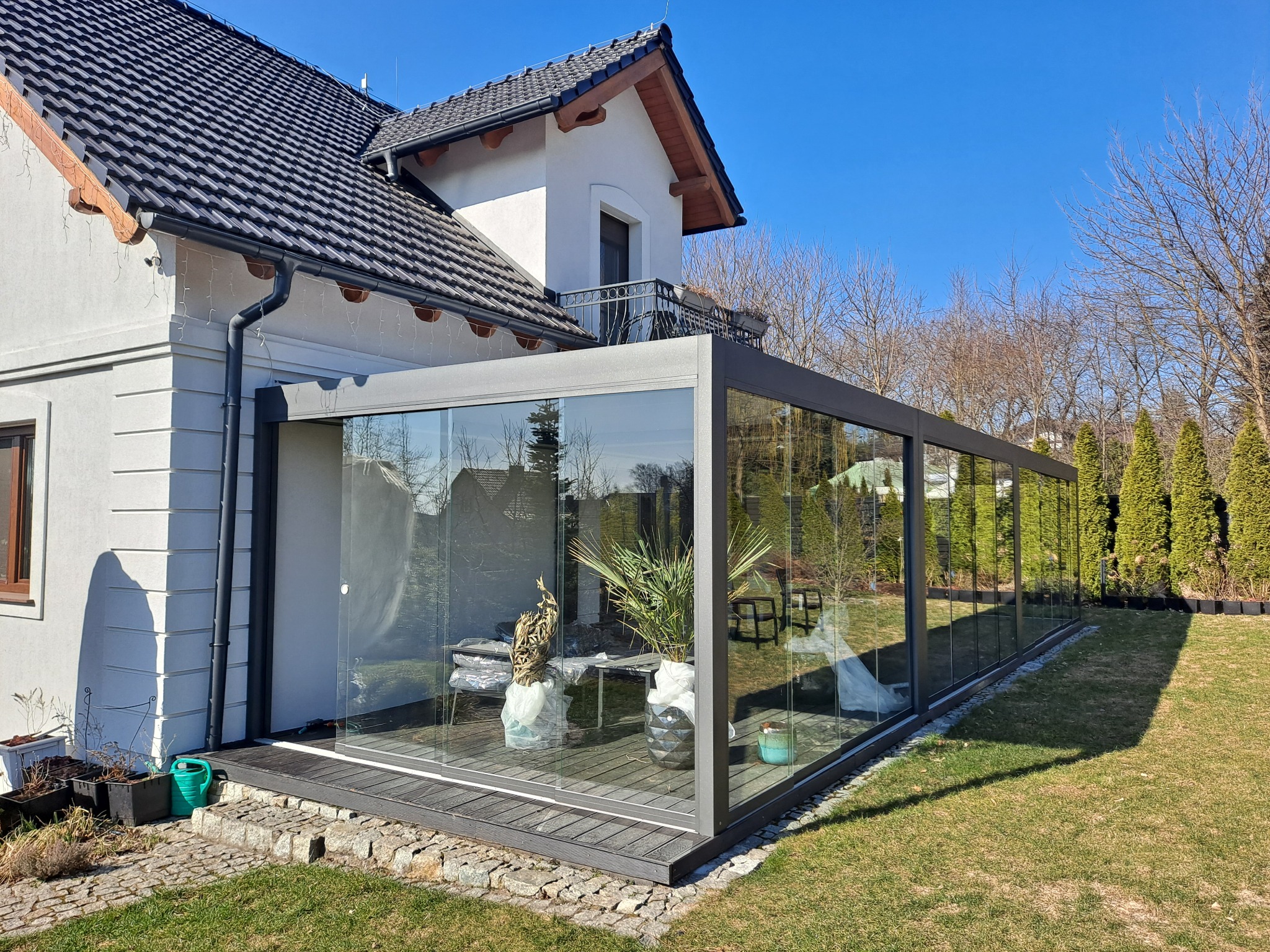 Szklana zabudowa tarasu z metalową ramą, przylegająca do białego domu z ciemnym dachem. Wewnątrz rośliny doniczkowe i meble ogrodowe. Słoneczny dzień.