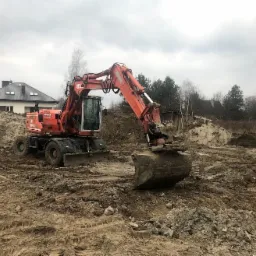 Pomarańczowa koparka kołowa Atlas na placu budowy, z wykopanym dołem i stertą ziemi w tle, widoczny dom jednorodzinny z szarą dachówką.