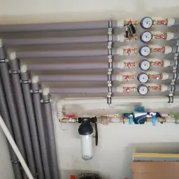 Rzędy rur hydraulicznych z izolacją termiczną i licznikami, przymocowane do ściany za pomocą metalowych obejm.