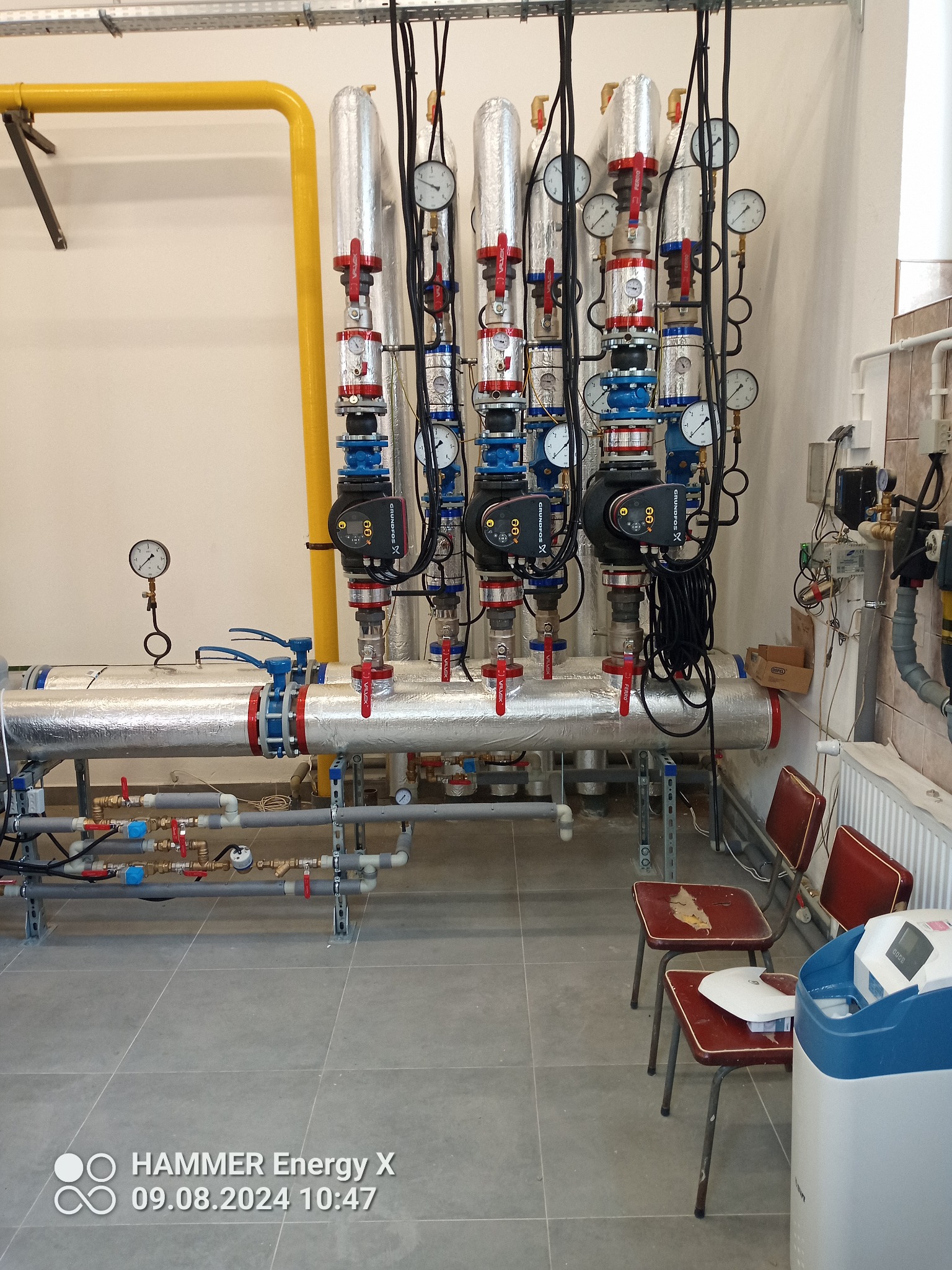 Rozbudowana instalacja grzewcza z rurami w izolacji, manometrami i pompami obiegowymi, zamontowana w pomieszczeniu technicznym. Widoczne liczne zawory i przyłącza.
