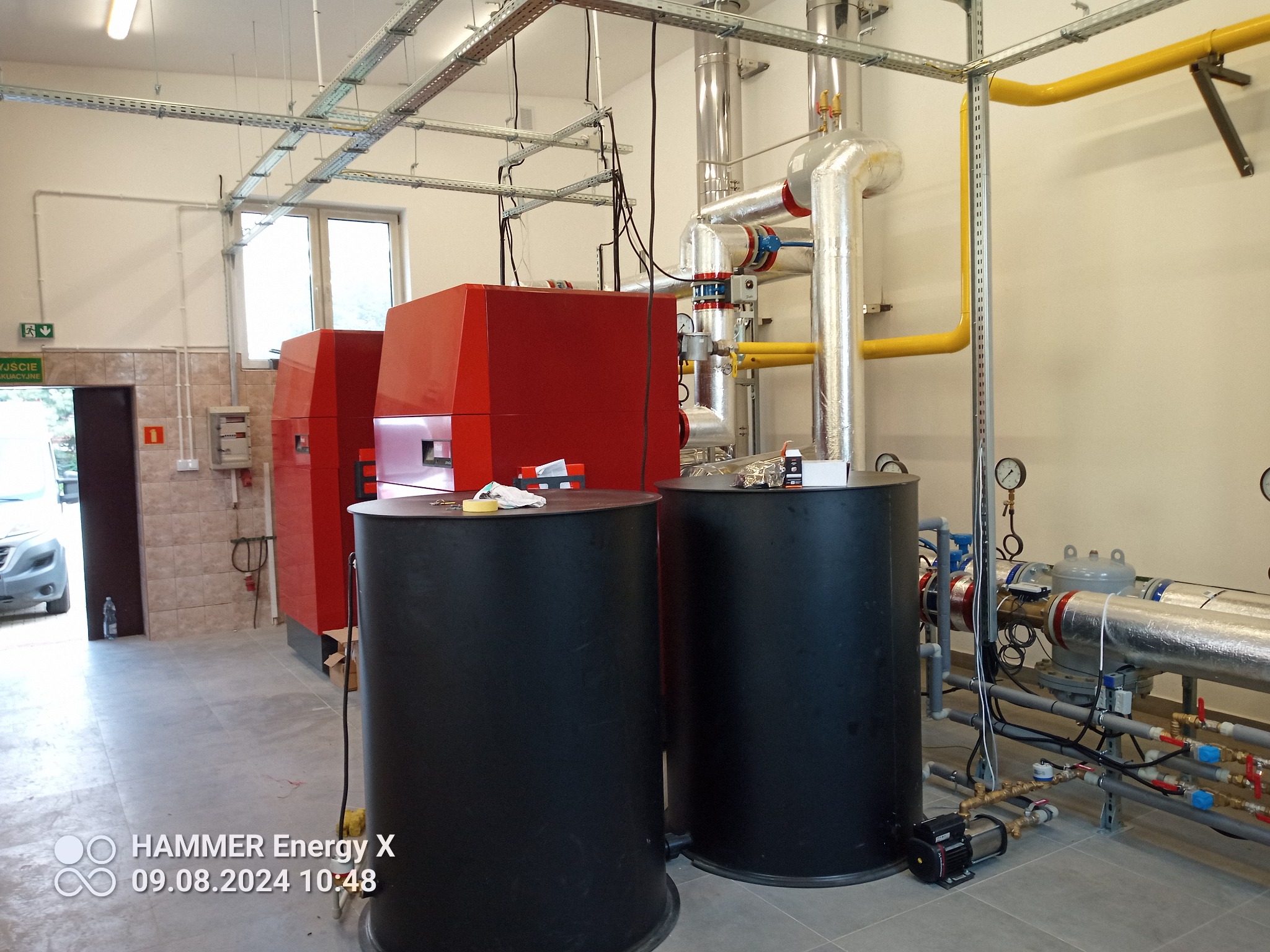 Nowoczesna kotłownia z czerwonymi kotłami i czarnymi zbiornikami buforowymi. Widoczne rury, zawory i instalacje. Oświetlenie sufitowe i okno w tle.