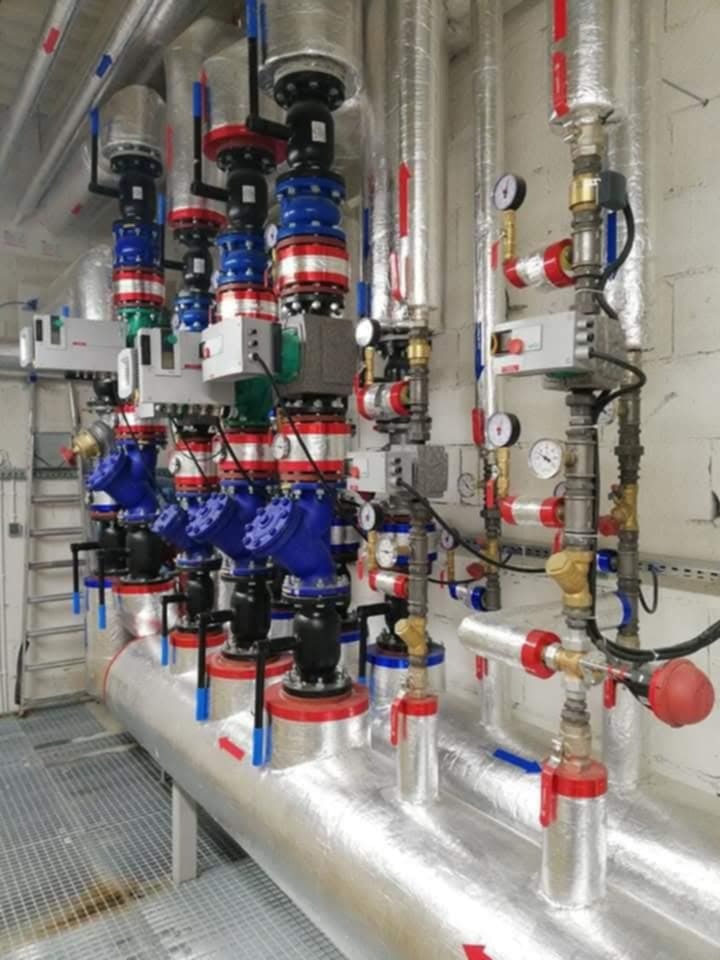 Złożona instalacja przemysłowa z rur, zaworów i manometrów, owinięta srebrną izolacją, z elementami w kolorach niebieskim, czerwonym i czarnym, na tle białej ściany.