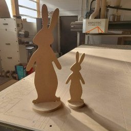 Szymon Babik - Dwie drewniane figury królików, większa i mniejsza, stoją na stole roboczym frezarki CNC marki Infotec, w tle widoczna maszyna i okno.