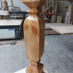 Szymon Babik - Toczone drewniane tralki schodowe z widocznym pęknięciem, w tle maszyna CNC w warsztacie stolarskim.