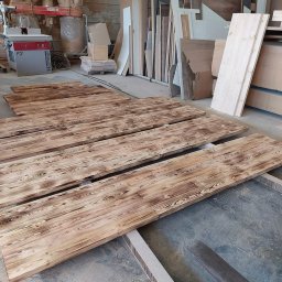 Szymon Babik - Opalane deski drewniane z wyraźnym usłojeniem, leżące na podłodze warsztatu, w tle maszyny stolarskie i składowane płyty drewniane.