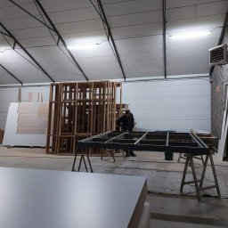 Szymon Babik - Warsztat stolarski z widocznym szkieletem konstrukcji szafy lub mebla na wymiar, stosem płyt meblowych i pracującym stolarzem w tle.