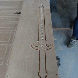 Szymon Babik - Płyta z jasnego drewna z wyfrezowanym wzorem miecza, widoczne ślady obróbki i fragment dłoni w tle.