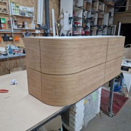 Szymon Babik - Wykończona, fornirowana szafka z zaokrąglonymi frontami, stojąca w warsztacie stolarskim na stole roboczym, otoczona narzędziami i materiałami.