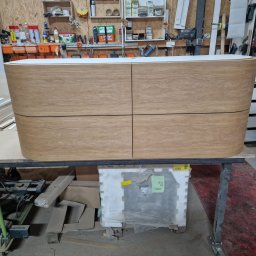 Szymon Babik - Wykończona komoda z czterema szufladami z jasnego drewna o zaokrąglonych krawędziach, ustawiona na metalowym stole w warsztacie stolarskim. W tle widoczne półki z narzędziami i materiałami.