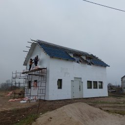 Szymon Babik - Budowa domu: Dwóch pracowników na rusztowaniu montuje izolację termiczną na ścianie zewnętrznej budynku z częściowo położonym dachem z niebieskiej dachówki. Na pierwszym planie widoczny duży stos...