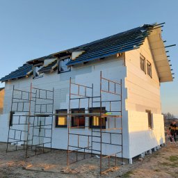 Szymon Babik - Dom w trakcie ocieplania styropianem, widoczne rusztowania i częściowo położone pokrycie dachu z niebieską membraną, w tle osoba z drabiną.