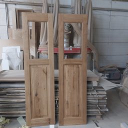 Szymon Babik - Dwie surowe, drewniane ramy drzwiowe z szybami, stojące w warsztacie stolarskim na tle maszyny do obróbki drewna i stosu desek.