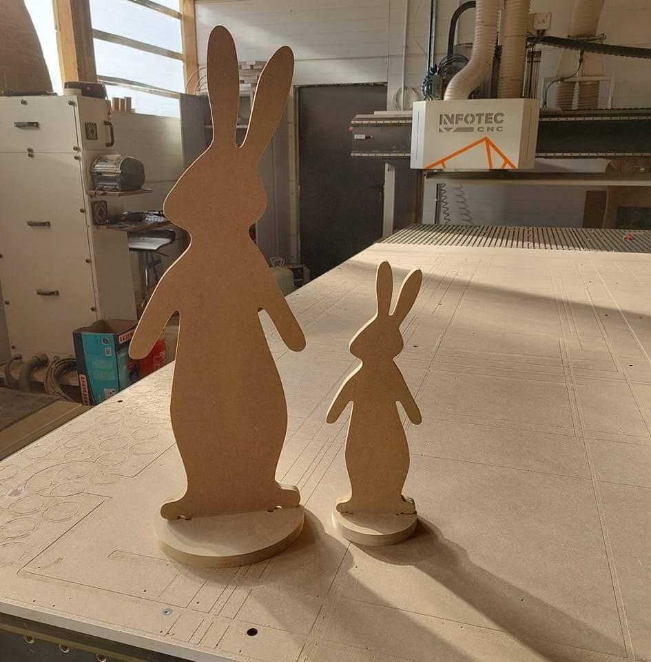 Dwie drewniane figury królików, większa i mniejsza, stoją na stole roboczym frezarki CNC marki Infotec, w tle widoczna maszyna i okno.