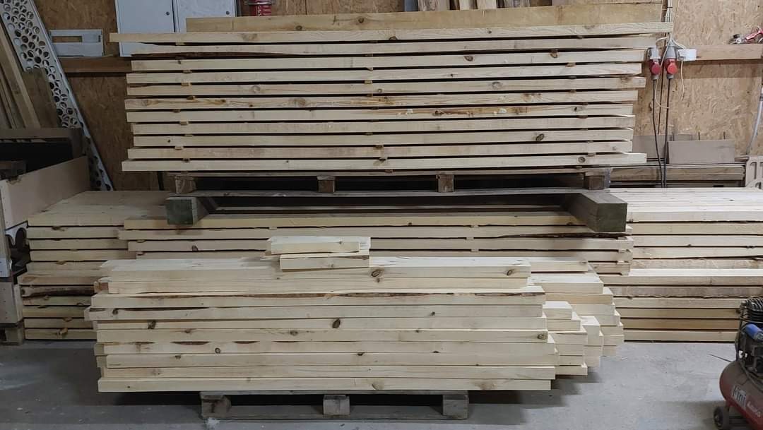 Stosy świeżego, jasnego drewna ułożone na paletach w warsztacie, widoczne sęki i naturalny rysunek drewna, w tle narzędzia i instalacje elektryczne.