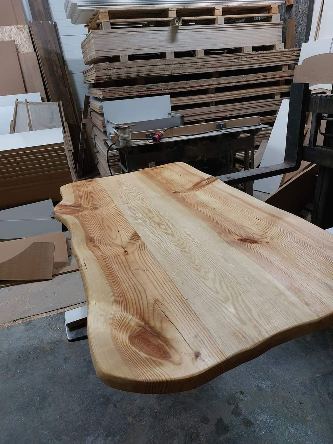 Blat stołu z surowego drewna o nieregularnych krawędziach, widoczne słoje i naturalne niedoskonałości, w tle warsztat stolarski z paletami i płytami.