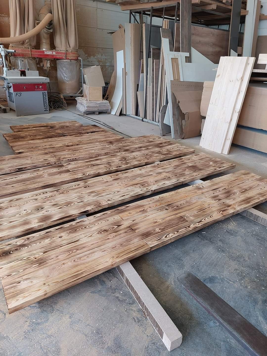 Opalane deski drewniane z wyraźnym usłojeniem, leżące na podłodze warsztatu, w tle maszyny stolarskie i składowane płyty drewniane.