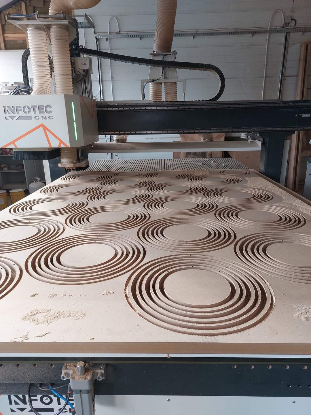 Frezarka CNC wycina wzór koncentrycznych okręgów w płycie drewnianej, widoczne ślady pyłu po obróbce, maszyna marki Infotec CNC.