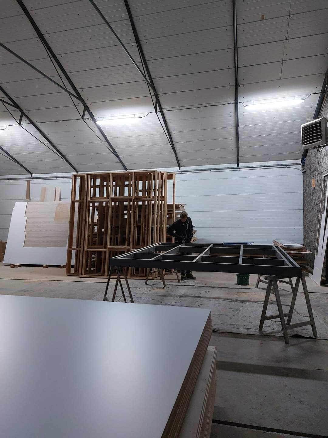 Warsztat stolarski z widocznym szkieletem konstrukcji szafy lub mebla na wymiar, stosem płyt meblowych i pracującym stolarzem w tle.