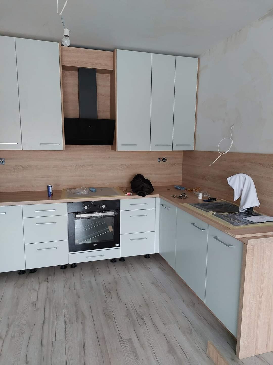 Montaż jasnej kuchni: białe fronty szafek dolnych i górnych, drewnopodobny blat i panel ścienny, czarny okap, płyta indukcyjna i piekarnik w zabudowie, widoczne elementy montażowe i narzędzia.