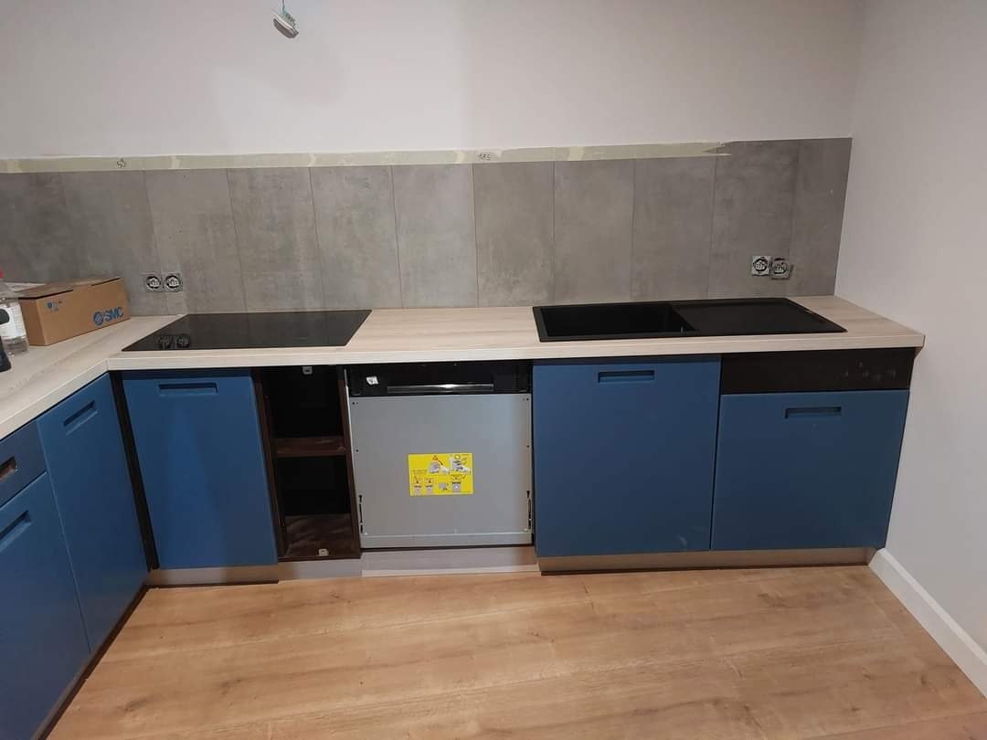 Niebieskie szafki kuchenne z blatem w kolorze jasnego drewna, płyta indukcyjna i zlewozmywak wpuszczone w blat, szare płytki na ścianie, częściowo zamontowana zmywarka, podłoga z paneli imitujących...