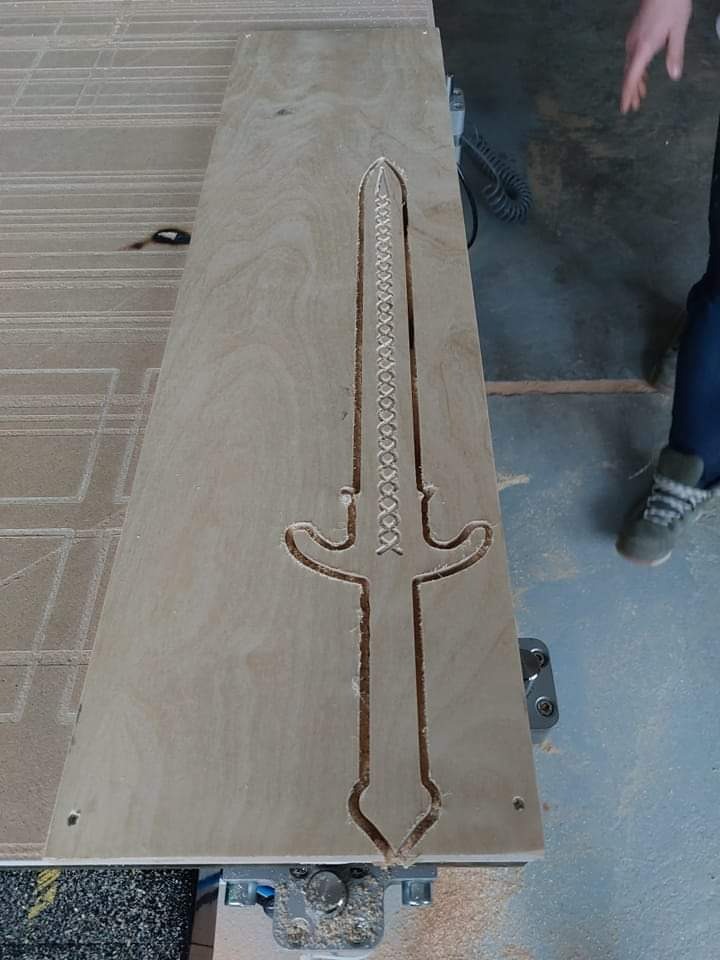 Płyta z jasnego drewna z wyfrezowanym wzorem miecza, widoczne ślady obróbki i fragment dłoni w tle.