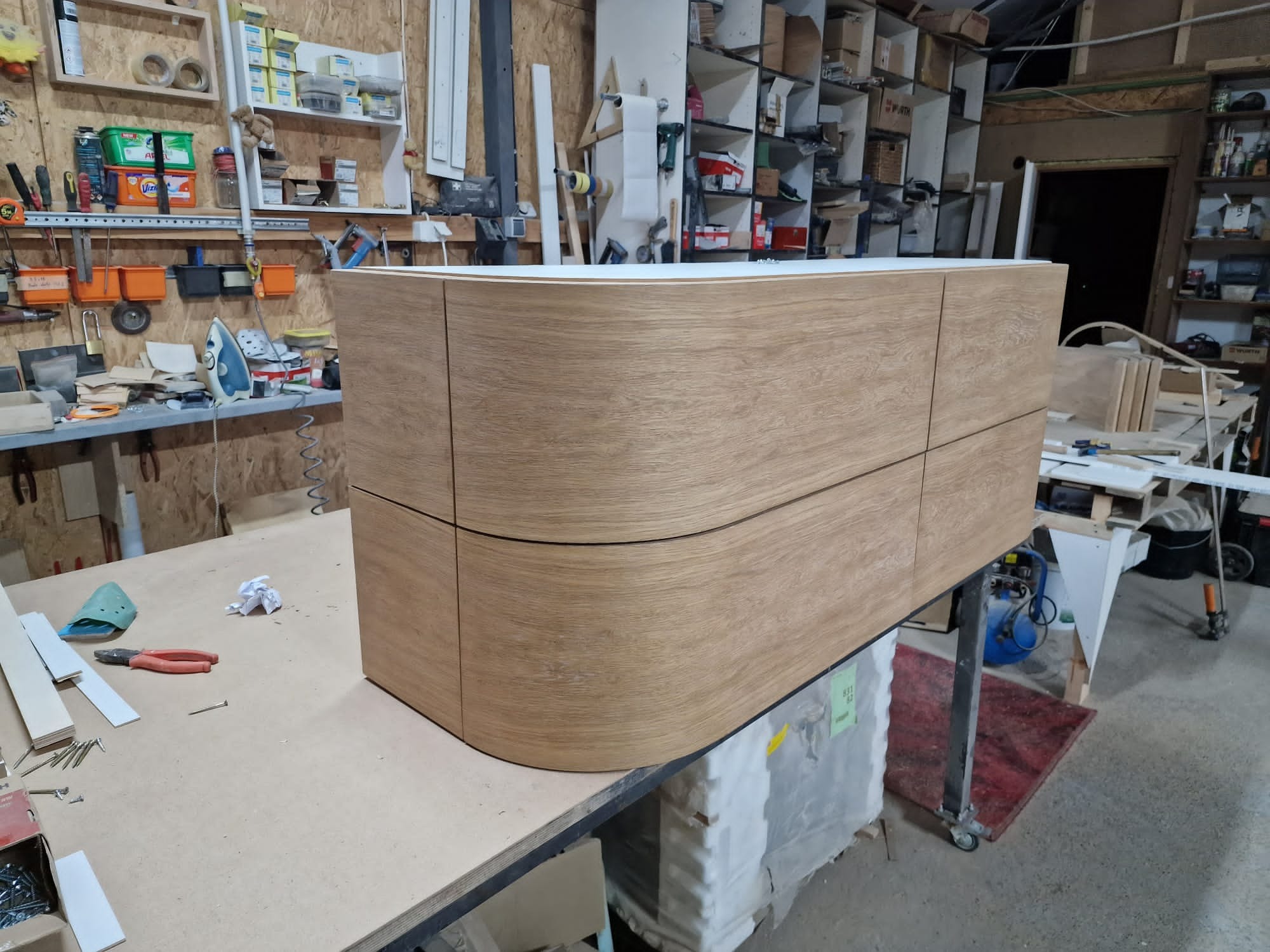 Wykończona, fornirowana szafka z zaokrąglonymi frontami, stojąca w warsztacie stolarskim na stole roboczym, otoczona narzędziami i materiałami.