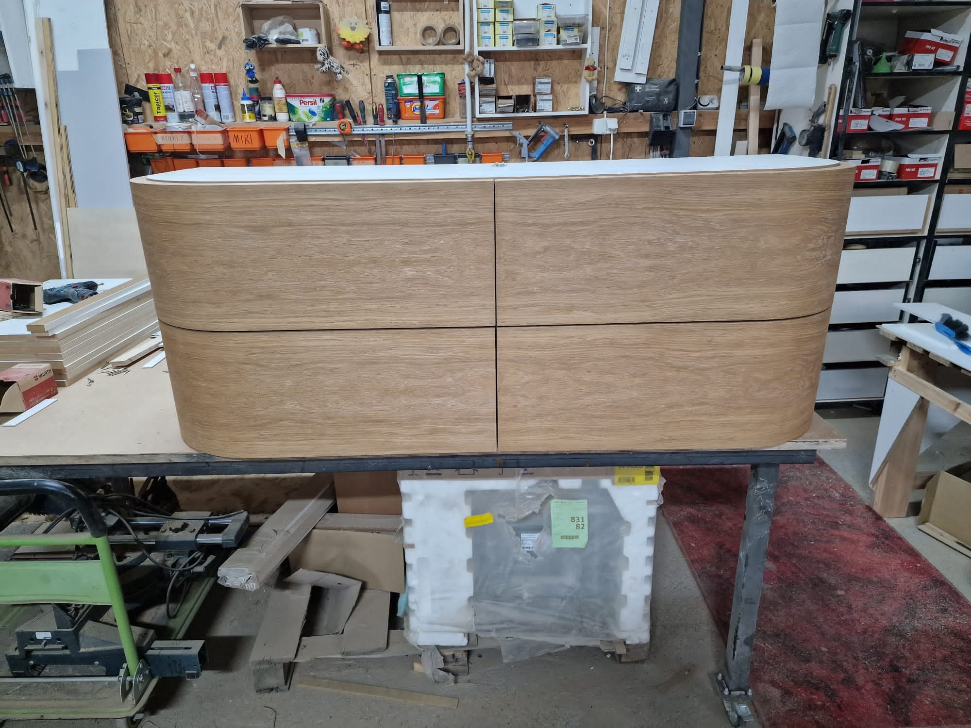 Wykończona komoda z czterema szufladami z jasnego drewna o zaokrąglonych krawędziach, ustawiona na metalowym stole w warsztacie stolarskim. W tle widoczne półki z narzędziami i materiałami.