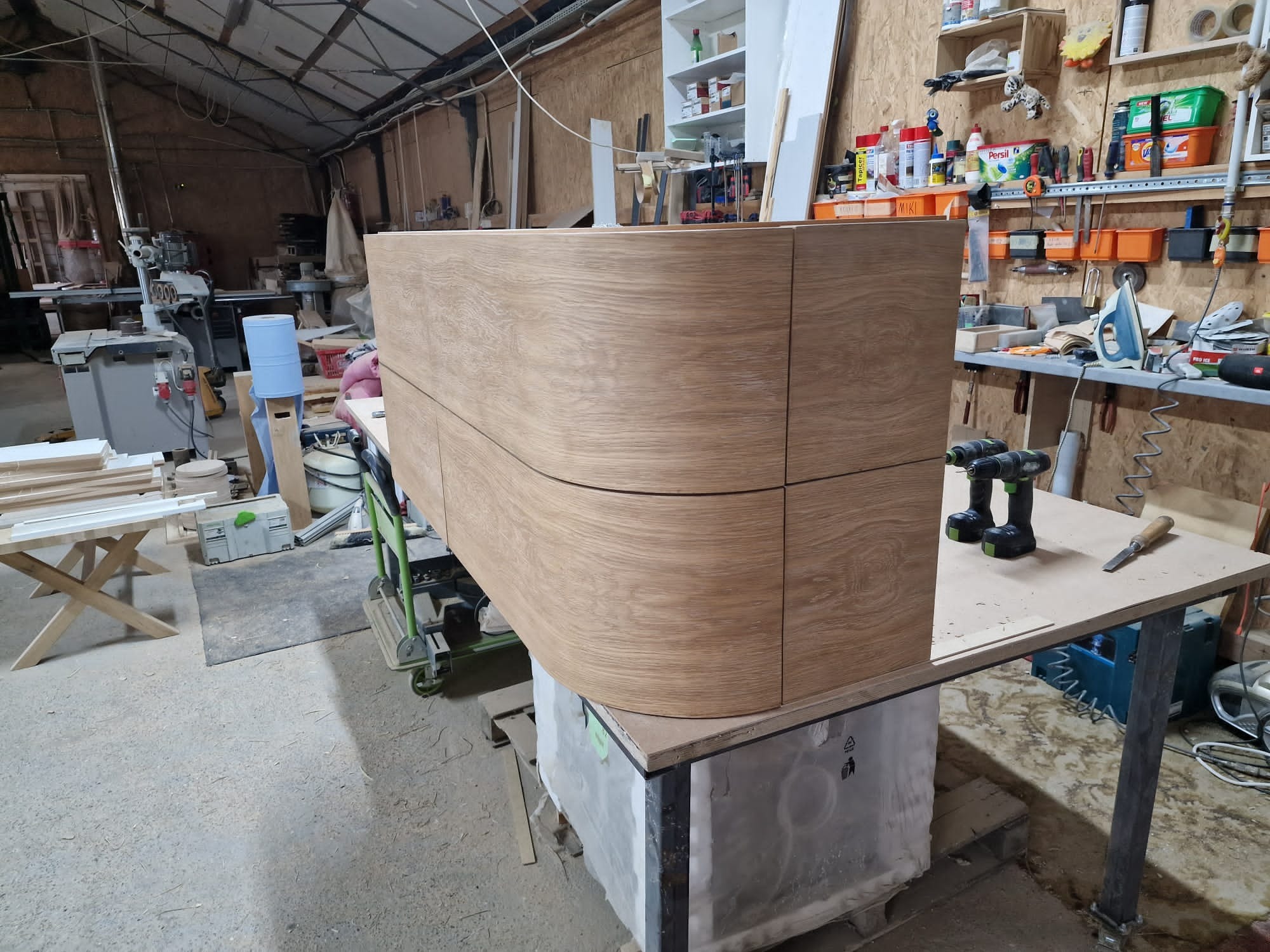 Częściowo zmontowana szafka z zaokrąglonymi frontami z jasnego drewna dębowego w warsztacie, widoczne wkrętarki akumulatorowe i dłuto na stole roboczym.