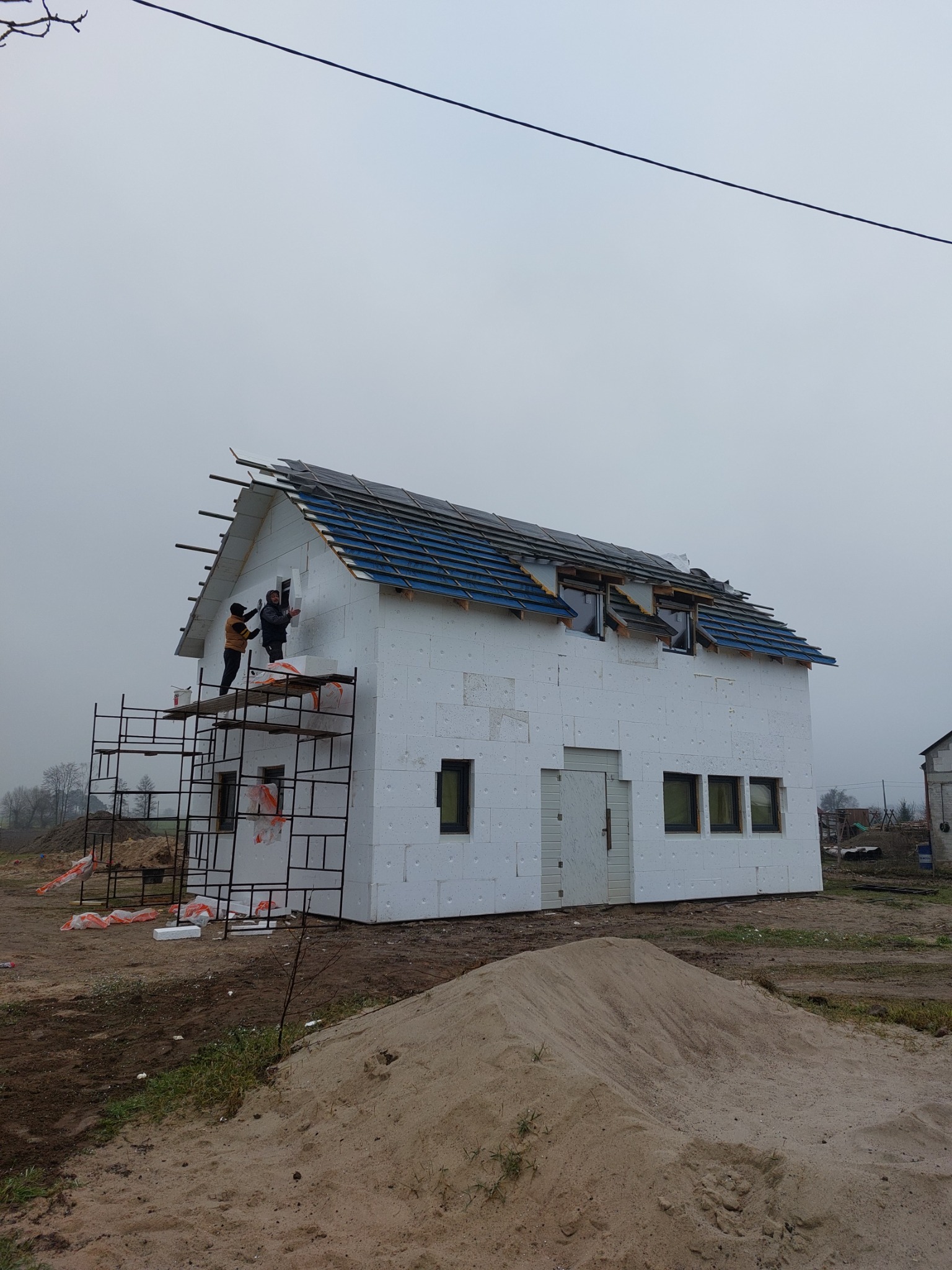 Budowa domu: Dwóch pracowników na rusztowaniu montuje izolację termiczną na ścianie zewnętrznej budynku z częściowo położonym dachem z niebieskiej dachówki. Na pierwszym planie widoczny duży stos...