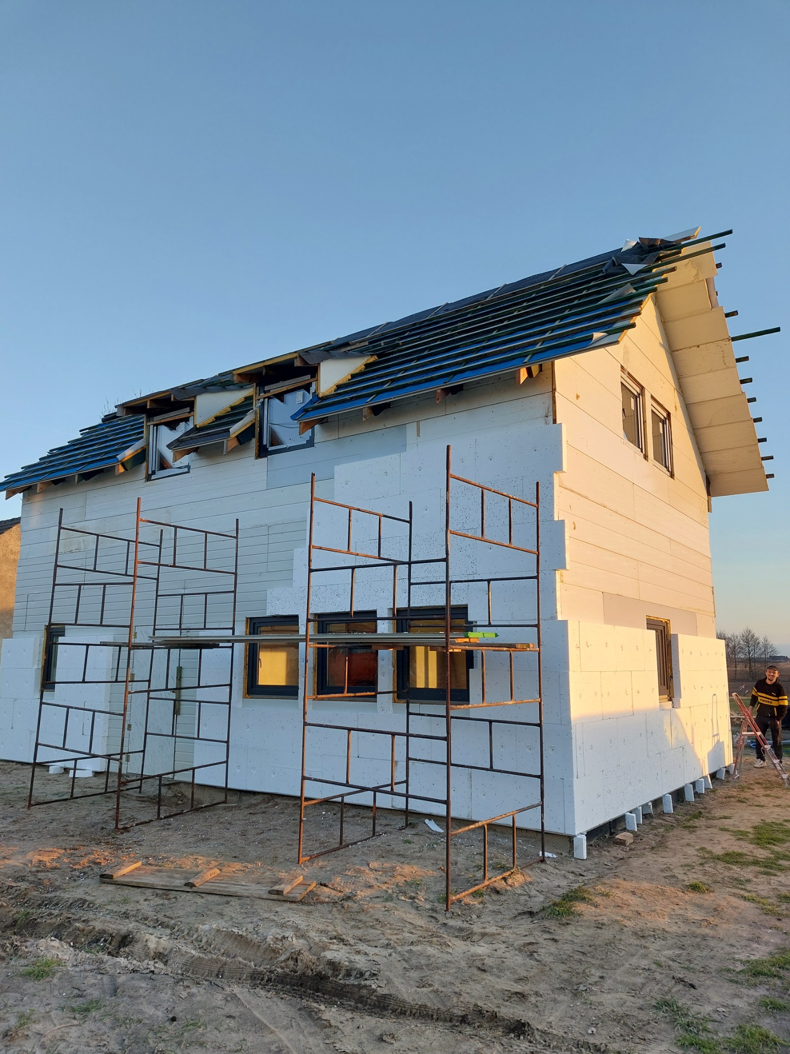 Dom w trakcie ocieplania styropianem, widoczne rusztowania i częściowo położone pokrycie dachu z niebieską membraną, w tle osoba z drabiną.