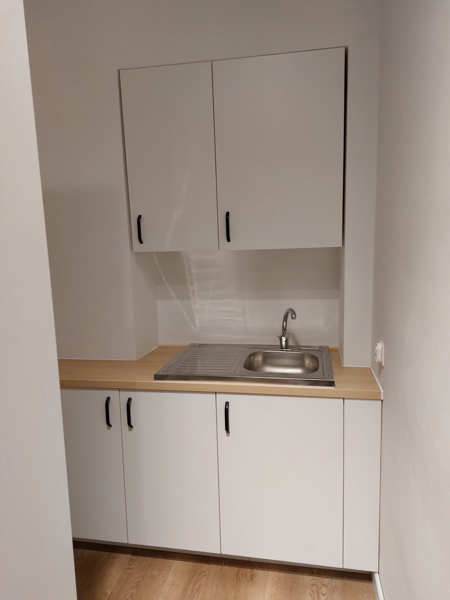 Nowoczesna, minimalistyczna kuchnia z białymi szafkami i drewnianym blatem, zlew ze stali nierdzewnej i bateria, widoczny fragment podłogi z paneli.
