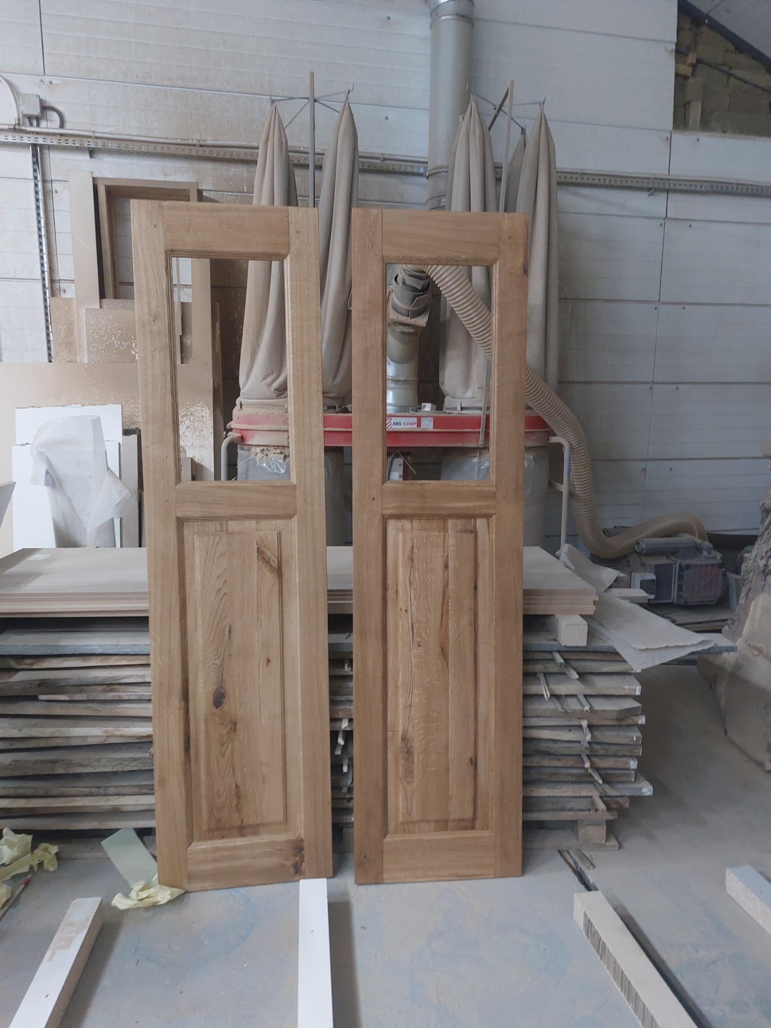 Dwie surowe, drewniane ramy drzwiowe z szybami, stojące w warsztacie stolarskim na tle maszyny do obróbki drewna i stosu desek.