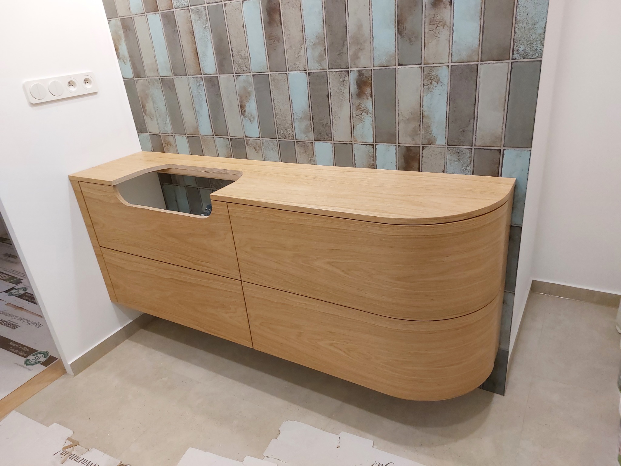 Drewniana szafka łazienkowa pod umywalkę z zaokrąglonymi bokami i czterema szufladami, zamontowana na ścianie z dekoracyjnymi, pionowymi płytkami w odcieniach brązu i błękitu. Na podłodze widoczne...