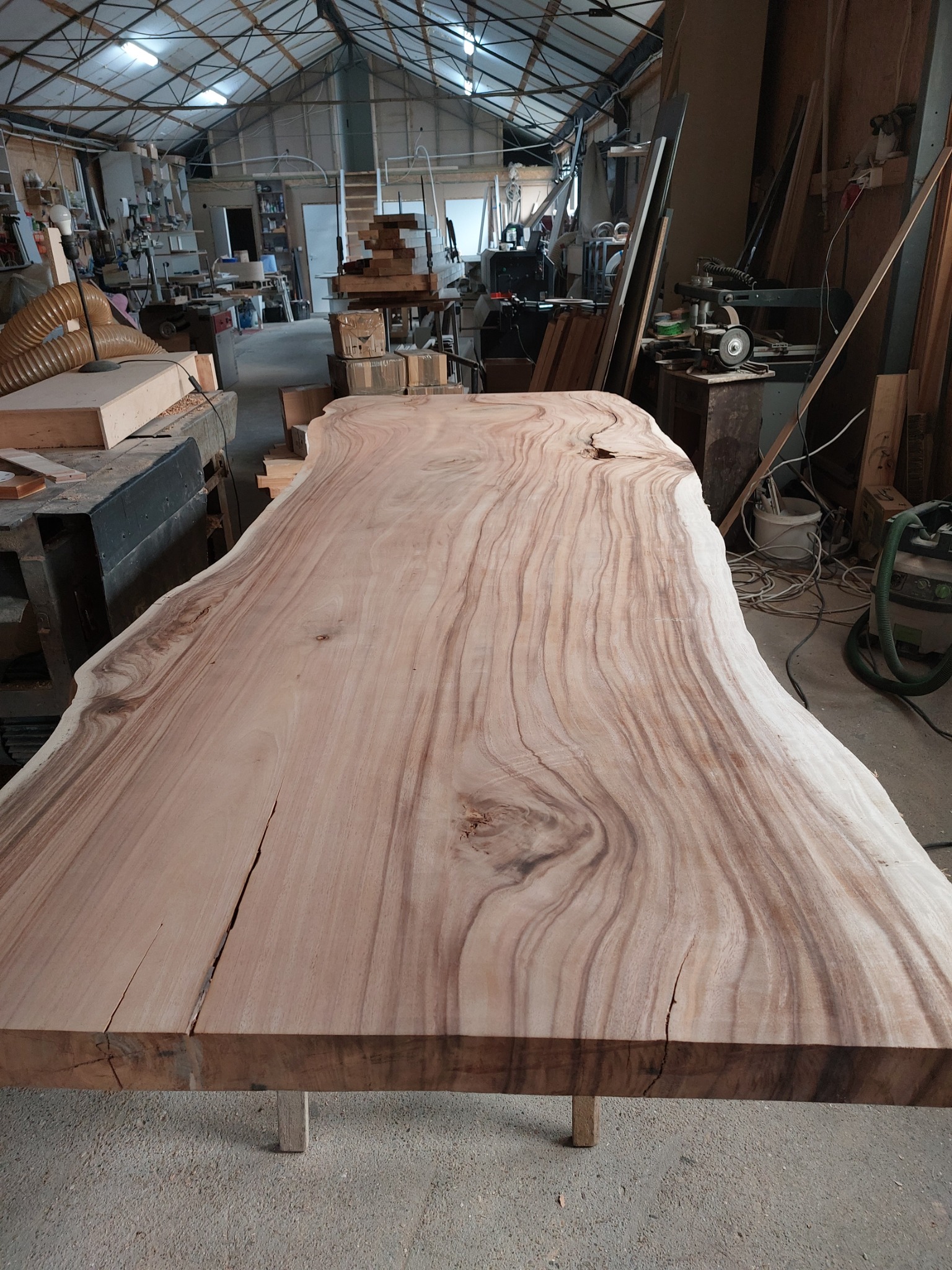 Surowy blat stołu z litego drewna o nieregularnym kształcie i wyraźnym usłojeniu, umieszczony w warsztacie stolarskim z widocznymi narzędziami i materiałami.