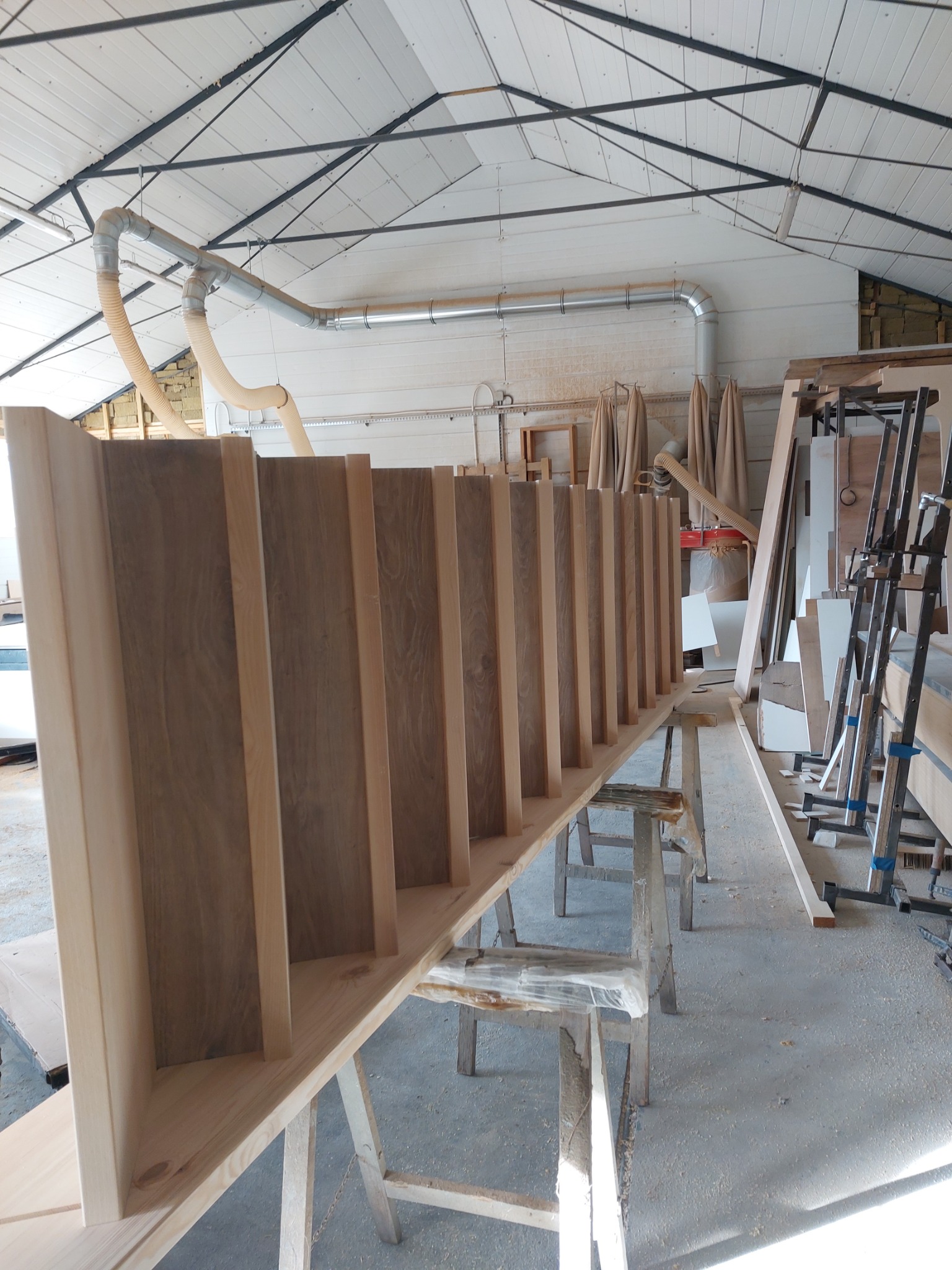 Element szafy na wymiar w trakcie produkcji w warsztacie stolarskim, widoczne drewniane listwy i elementy konstrukcyjne, system odciągów pyłu pod sufitem.