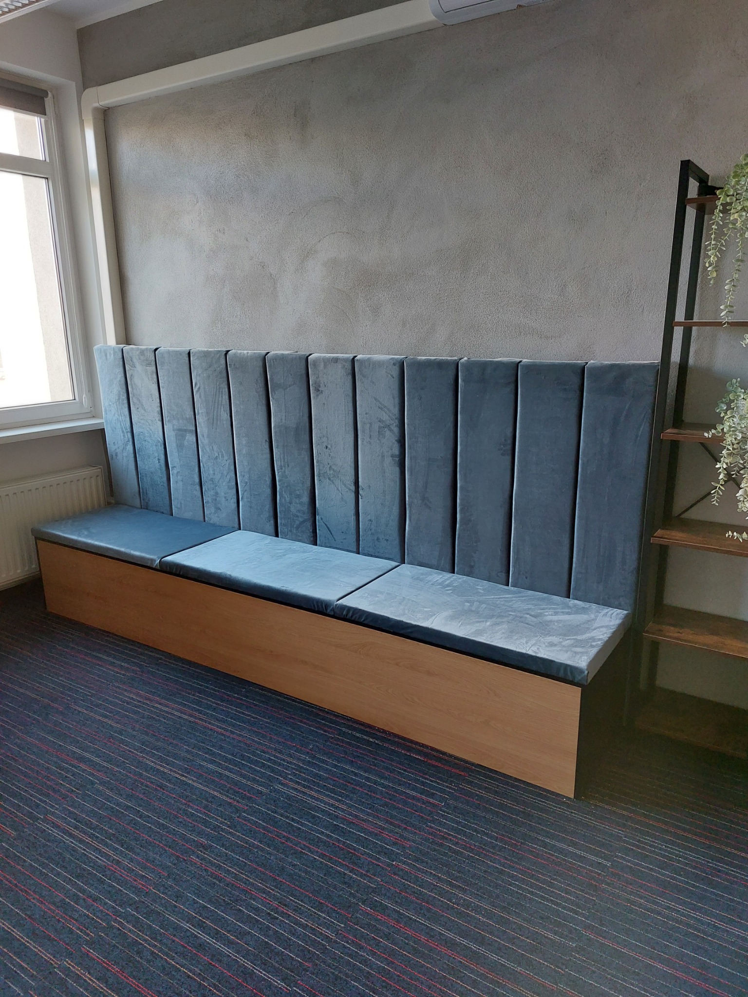 Długa, tapicerowana ławka z wysokim, dzielonym na segmenty oparciem w kolorze szaroniebieskim, umieszczona na drewnianej podstawie z widoczną półką z roślinami w tle.