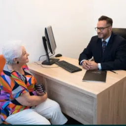 Doradca w garniturze rozmawia z klientką w kolorowej bluzce w jasnym biurze, na biurku komputer i czarny notatnik.