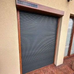 Zamknięta, grafitowa roleta zewnętrzna z brązową ramą i panelem solarnym u góry, zamontowana na elewacji budynku z terakotową podłogą.