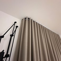 Bifam - Beżowe zasłony z równomiernymi fałdami zawieszone na czarnym karniszu, widoczne z dołu pod kątem, obok czarna lampa podłogowa.