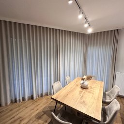 Bifam - Beżowe, marszczone zasłony do podłogi w nowoczesnym salonie z drewnianym stołem i krzesłami w odcieniach szarości, oświetlone reflektorami sufitowymi.
