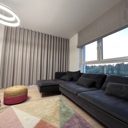 Bifam - Nowoczesny salon z szarą, falującą zasłoną, szarą roletą rzymską, ciemną sofą modułową i geometrycznym dywanem w pastelowych kolorach.
