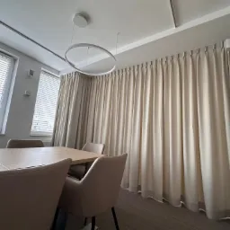 Nowoczesna sala konferencyjna z beżowymi zasłonami zakrywającymi całą ścianę, minimalistycznym okrągłym żyrandolem i dużym stołem otoczonym krzesłami w kolorze nude.