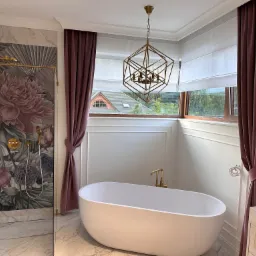 Elegancka łazienka z wolnostojącą wanną, dekoracyjną tapetą z motywem kwiatowym, nowoczesną lampą wiszącą i bordowymi zasłonami oraz roletami rzymskimi w oknie.